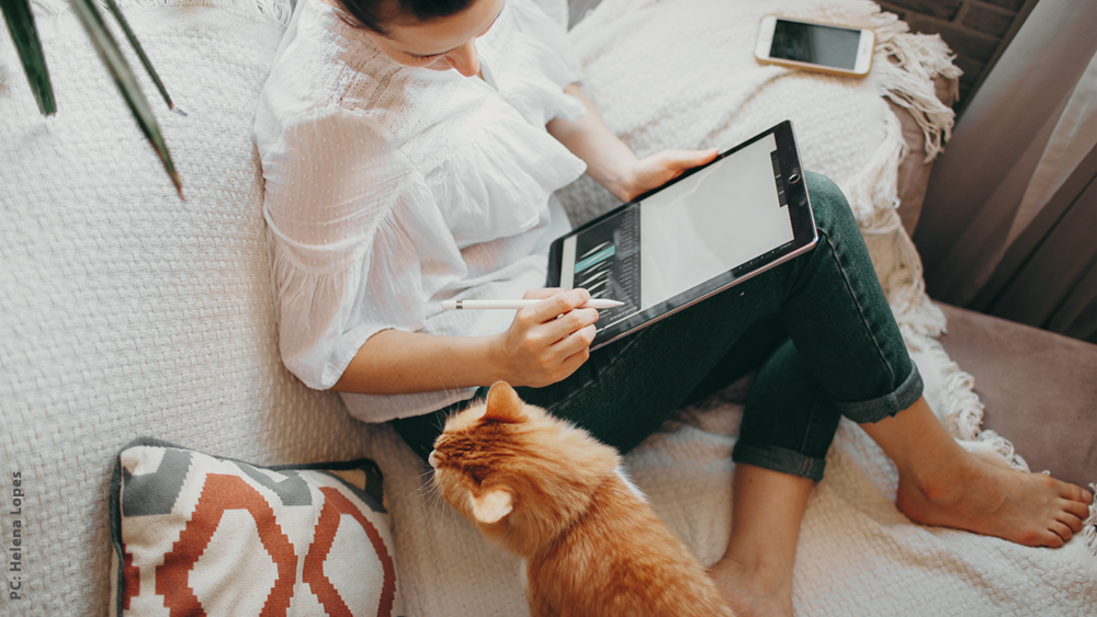 A woman and a ginger cat sitting on a bed, working from home, looking at an ipad